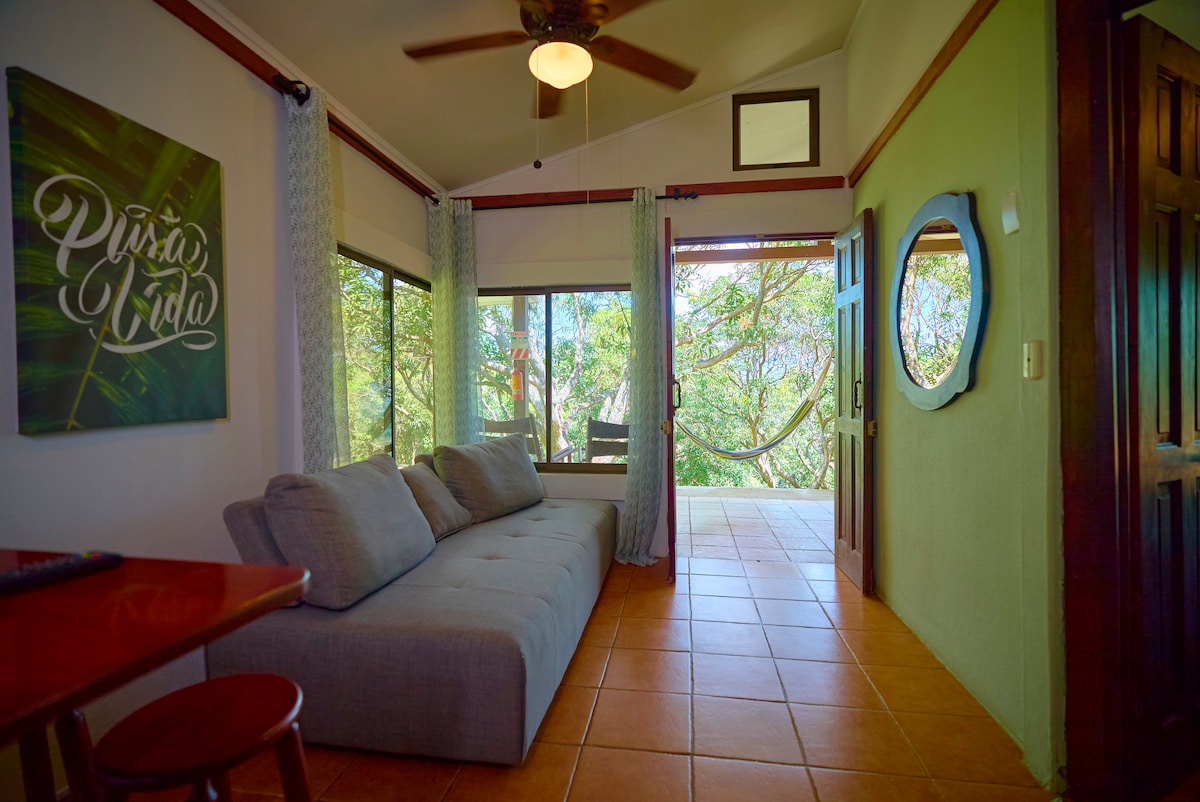 A cozy living area features a spacious gray sofa positioned near large windows, allowing natural light to brighten the space. Colorful art adorns the wall, and a small dining table complements the room. A wooden door leads to an outdoor patio area surrounded by trees.