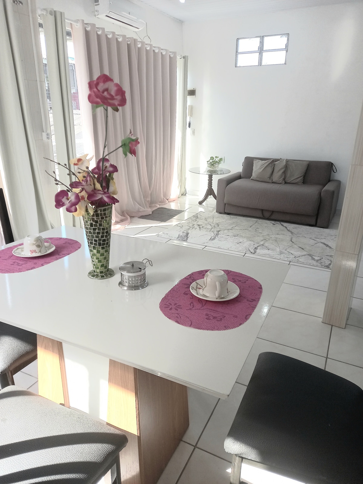 A dining area features a white table set with floral decorations and two tea cups, accompanied by black chairs. In the background, a comfortable gray sofa with cushions sits on a marble-patterned rug, illuminated by natural light from a nearby window.