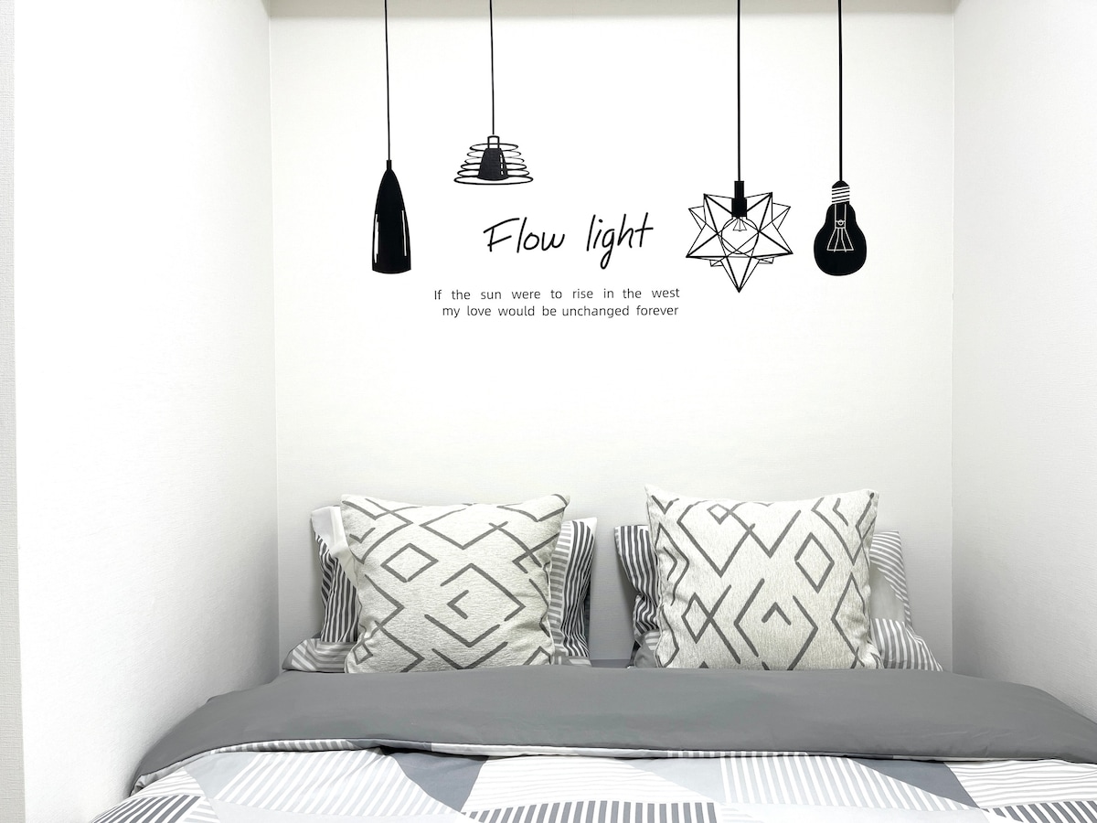 A sleeping area features a spacious bed with patterned gray and white bedding. Two decorative pillows are placed against the wall. Above, three stylish light fixtures hang elegantly, with a quote displayed on the wall, adding a distinctive touch.