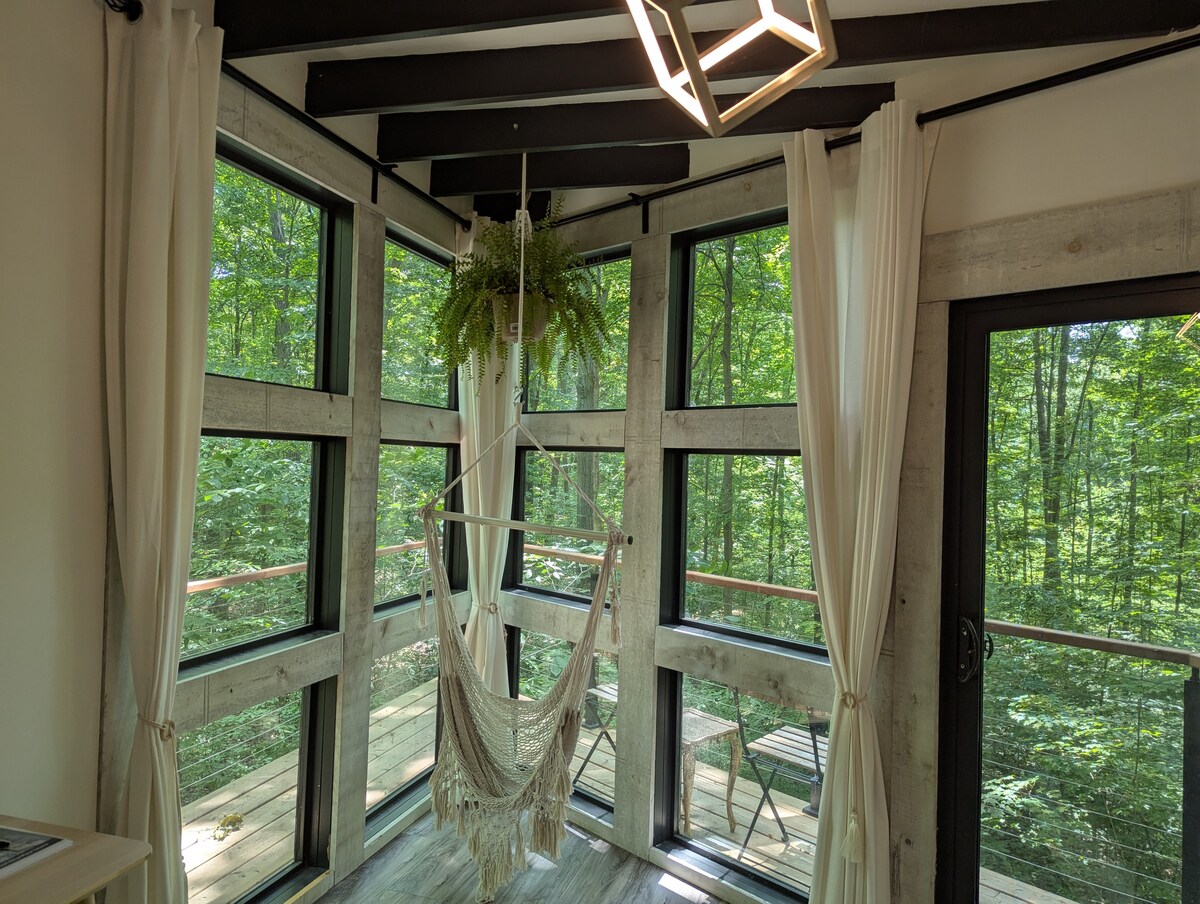 Large windows frame a serene view of the surrounding forest, allowing natural light to fill the room. A hammock hangs suspended, inviting relaxation. Sheer curtains soften the space, and a modern light fixture hangs from the ceiling, creating a harmonious blend of comfort and nature.
