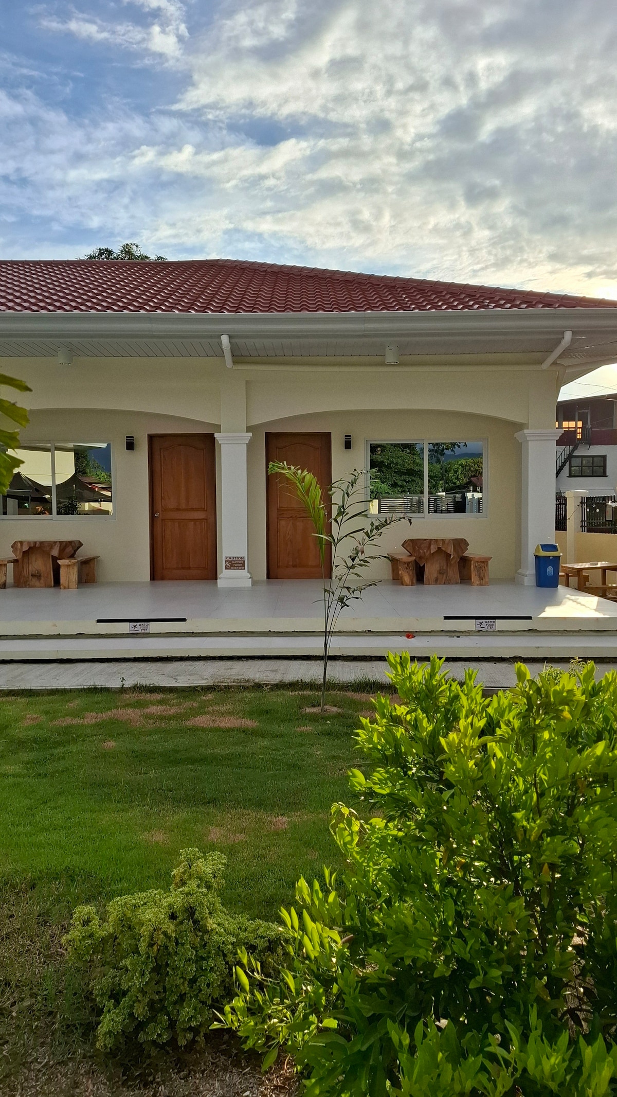 The exterior of the guest house features a porch with two wooden doors, flanked by large windows. Natural wooden furniture is arranged on the porch, surrounded by lush greenery. A well-maintained lawn enhances the inviting rural atmosphere of the space.