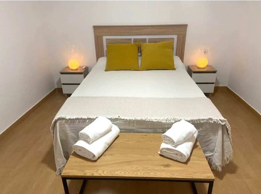 A spacious sleeping area features a large bed dressed with neutral linens and accented by two mustard pillows. Bedside tables with warm glowing lamps sit on either side. A wooden bench displays neatly rolled towels, enhancing the room's organized appearance.