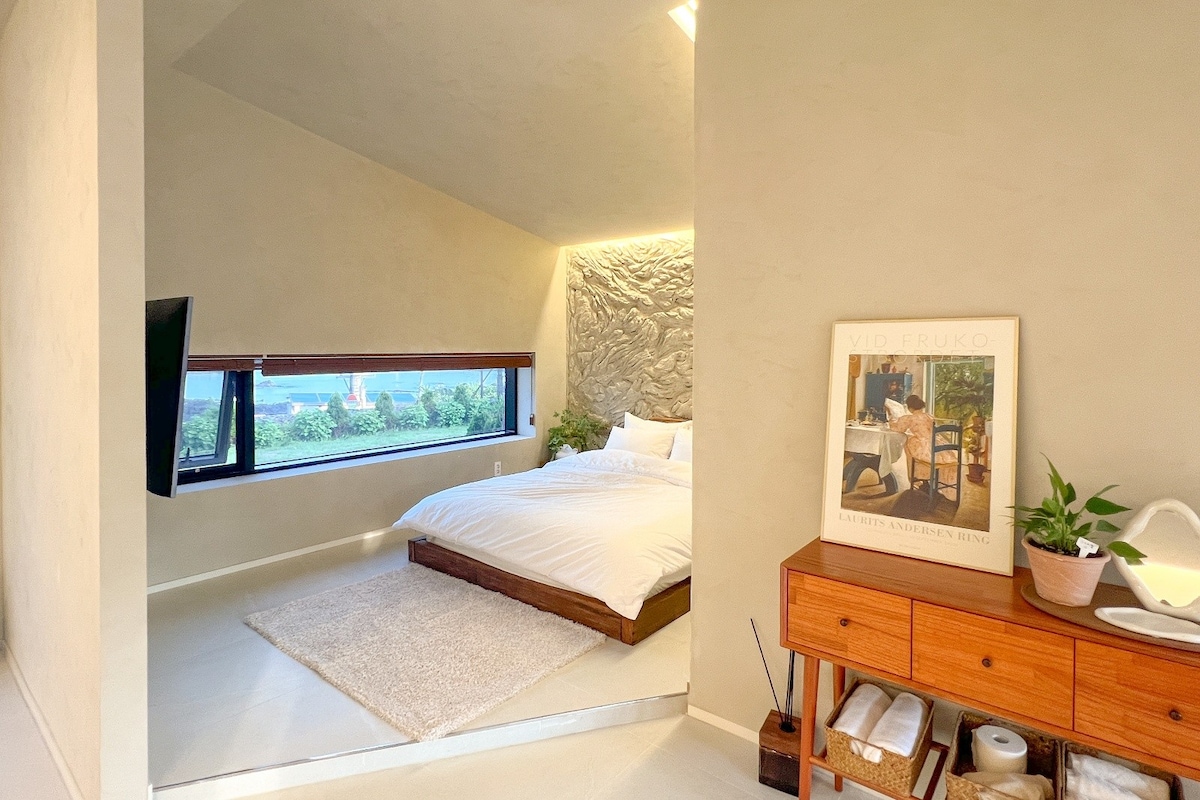 A cozy bedroom features a spacious bed with white linens against a backdrop of textured wall design. A large window provides a view of the outdoors, while a wooden dresser holds decorative elements, including a framed artwork and a small potted plant.