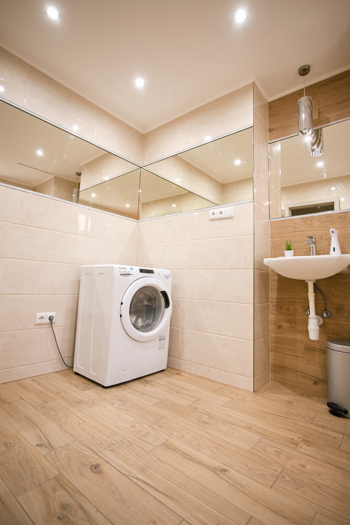 The spacious bathroom features modern fixtures, including a washing machine and a sink with a sleek design. Large mirrors reflect the light, enhancing the bright atmosphere. Neutral-toned tiles cover the walls and floor, creating a clean and functional space.