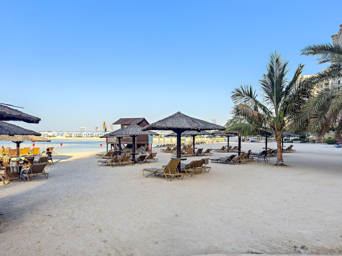 An inviting beach area features a smooth, sandy surface dotted with thatched-roof cabanas and lounge chairs. Palm trees provide shade, and the serene water is visible in the background, creating a relaxing atmosphere for unwinding by the shore.