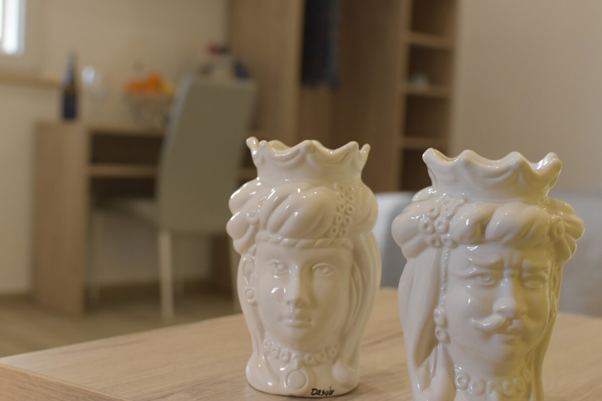 Two ceramic vases with floral designs are positioned on a wooden table. The background features a softly lit space with a work desk and a chair, providing an organized and calm environment.