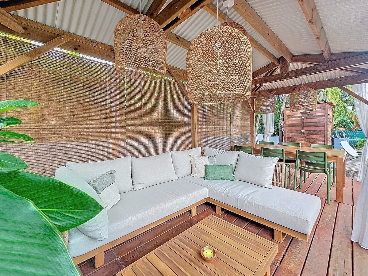A spacious lounge area is surrounded by natural bamboo screens, featuring a large sectional sofa adorned with light cushions. Decorative pendant lights hang overhead, while a wooden table serves as a centerpiece. A dining space with green chairs is visible in the background, blending outdoor and indoor elements.