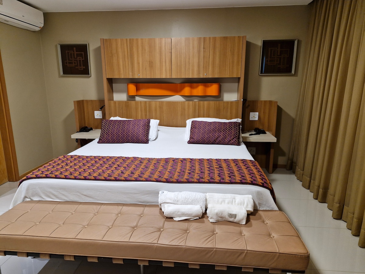 A spacious guest room features a king-size bed adorned with decorative pillows and a colorful throw. Wood accents on the headboard complement the neutral-toned walls. A bench sits at the foot of the bed, while bedside tables are equipped with reading lamps, enhancing the room's functionality.