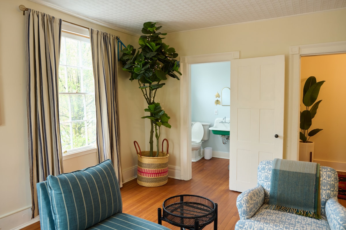 A light-filled living area displays a striped blue chair and a matching sofa. A large potted plant adds greenery, while the doorways lead to a bathroom and another room. Natural light illuminates the space through a large window with patterned curtains.