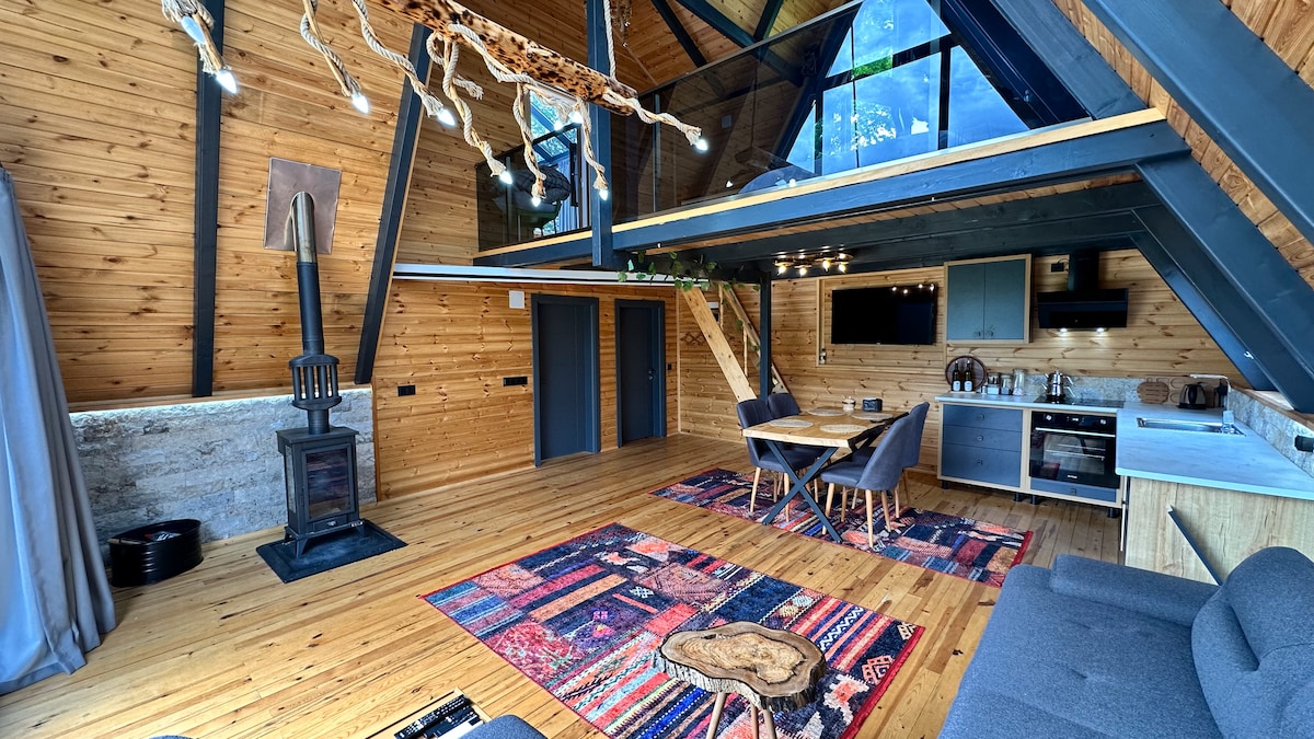 A spacious interior showcases an open layout with wooden walls and beams. A modern kitchen area is visible alongside a dining table and chairs. A rustic rug adds color to the wooden floor, while a cozy seating area is positioned near a decorative fireplace.