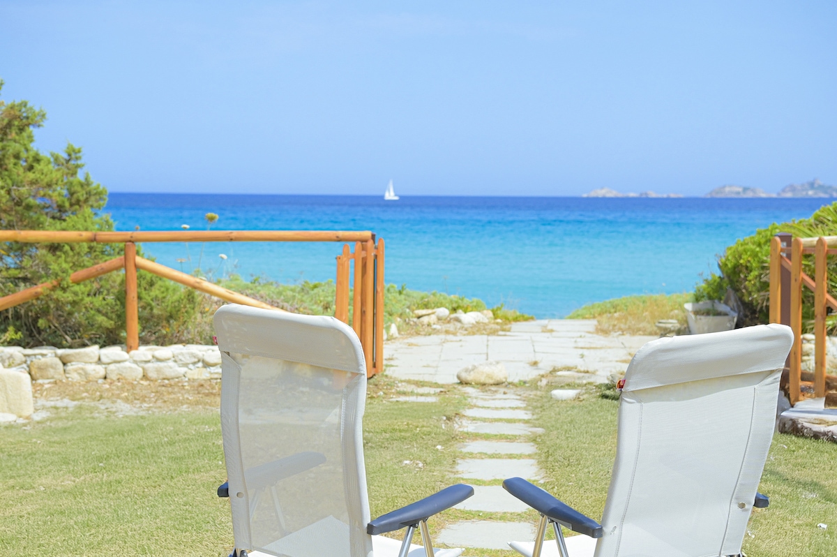 Two white lounge chairs are positioned on a grassy area, overlooking the calm turquoise sea. A path of stones leads from the chairs to a low wooden fence, with a sailboat visible in the distance under a clear blue sky.