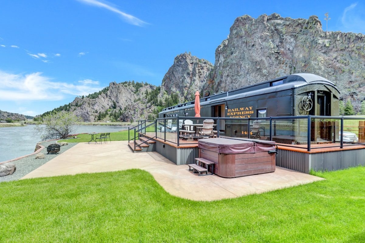 A historic train car is situated near the shoreline, complemented by a spacious deck featuring outdoor seating and a hot tub. Scenic rock formations rise in the background, while the nearby river enhances the natural landscape, creating a serene outdoor setting.