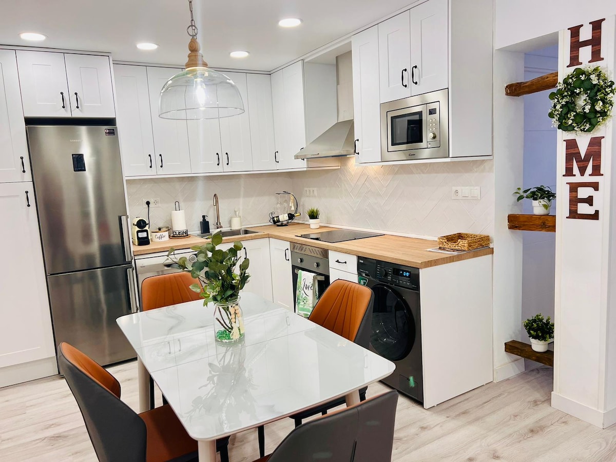 A modern kitchen is presented, featuring white cabinets and stainless steel appliances. A wooden countertop extends over the kitchen area, while a dining table is set for four. Decorative plants adorn the space, and a stylish light fixture hangs from the ceiling, enhancing the atmosphere.