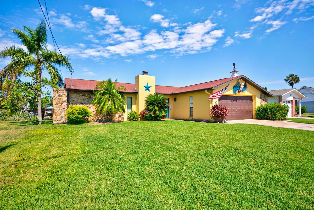 The exterior of a vibrant yellow home features a stone accent wall and a prominent star detail. Lush green grass surrounds the property, complemented by tropical palm trees. The driveway leads to a garage, and an American flag decorates the front, contributing to a welcoming presence.