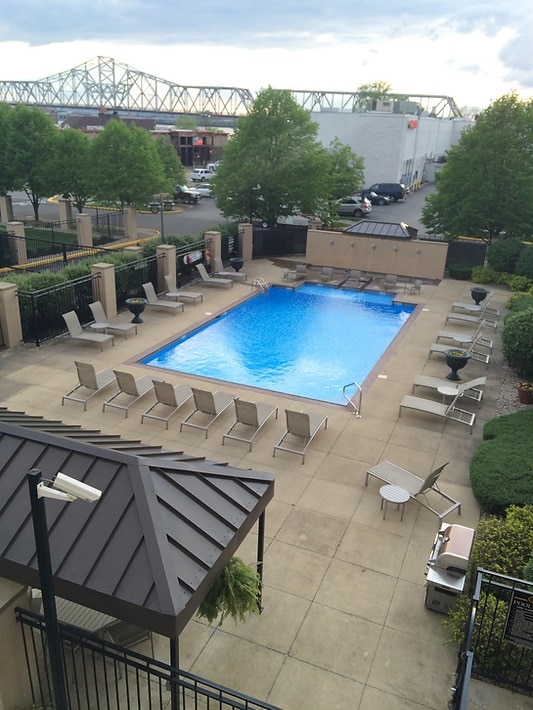 An outdoor heated pool is surrounded by a paved area with lounge chairs for relaxation. Lush trees and planters add greenery to the space. In the background, a bridge and buildings are visible, complementing the inviting atmosphere of the pool area.