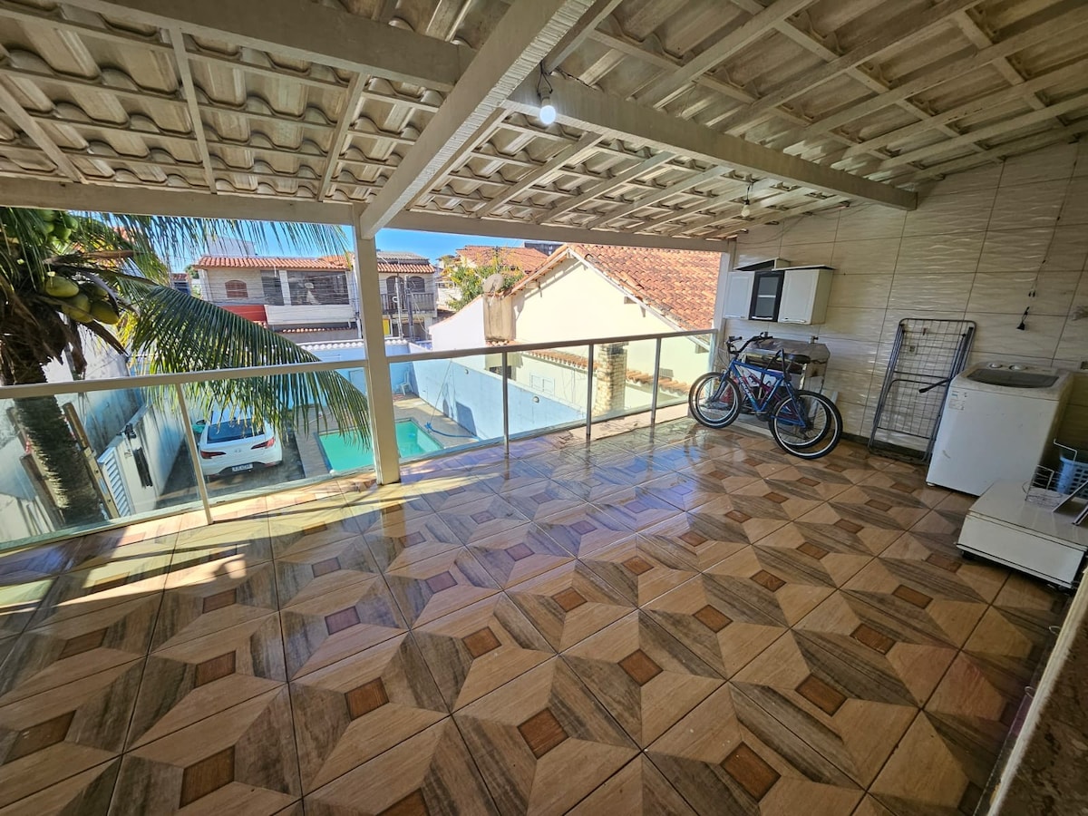 An open-air terrace is showcased, featuring a tiled floor and a covered ceiling. A washing machine and a drying rack are present, along with two bicycles resting against the wall. Sunlight streams in, creating a bright atmosphere.