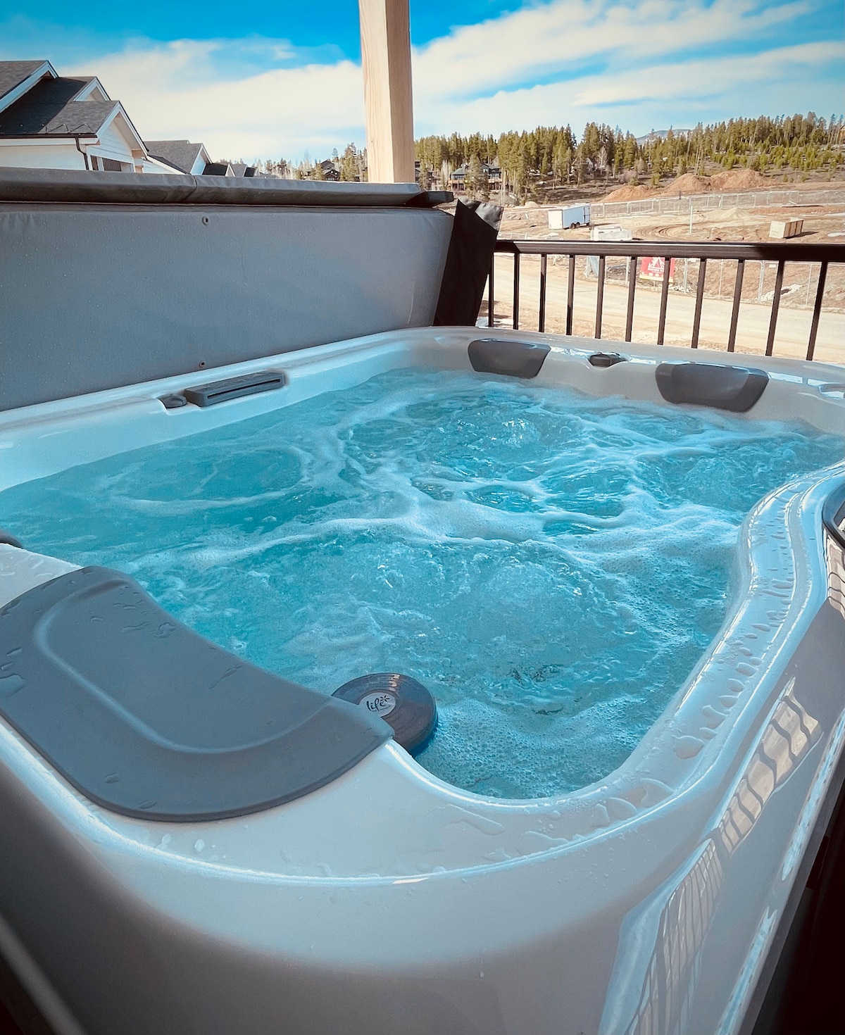 A private hot tub is filled with bubbling water, set against a backdrop of trees and distant hills. The tub's smooth edges and gray accents enhance its modern design. An outdoor railing frames the scene, providing a cozy and private atmosphere.