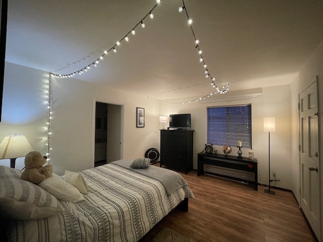A bedroom is presented with warm, ambient lighting from string lights overhead. A spacious bed with striped bedding is complemented by decorative pillows. A dark dresser stands nearby, next to a console table. A softly glowing lamp and a window with blinds provide additional lighting.