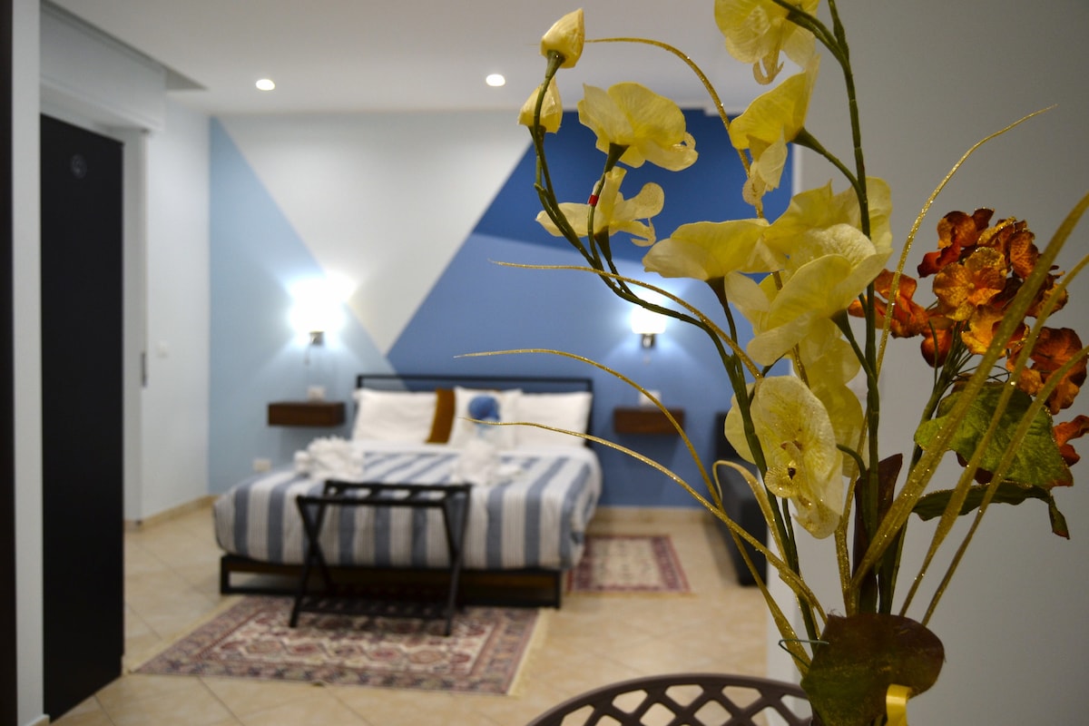 A spacious room is presented, featuring a striped bed with decorative pillows and bedside lamps. A floral arrangement is placed prominently in the foreground. A geometric accent wall adds visual interest, while warm-toned rugs enhance the floor space.