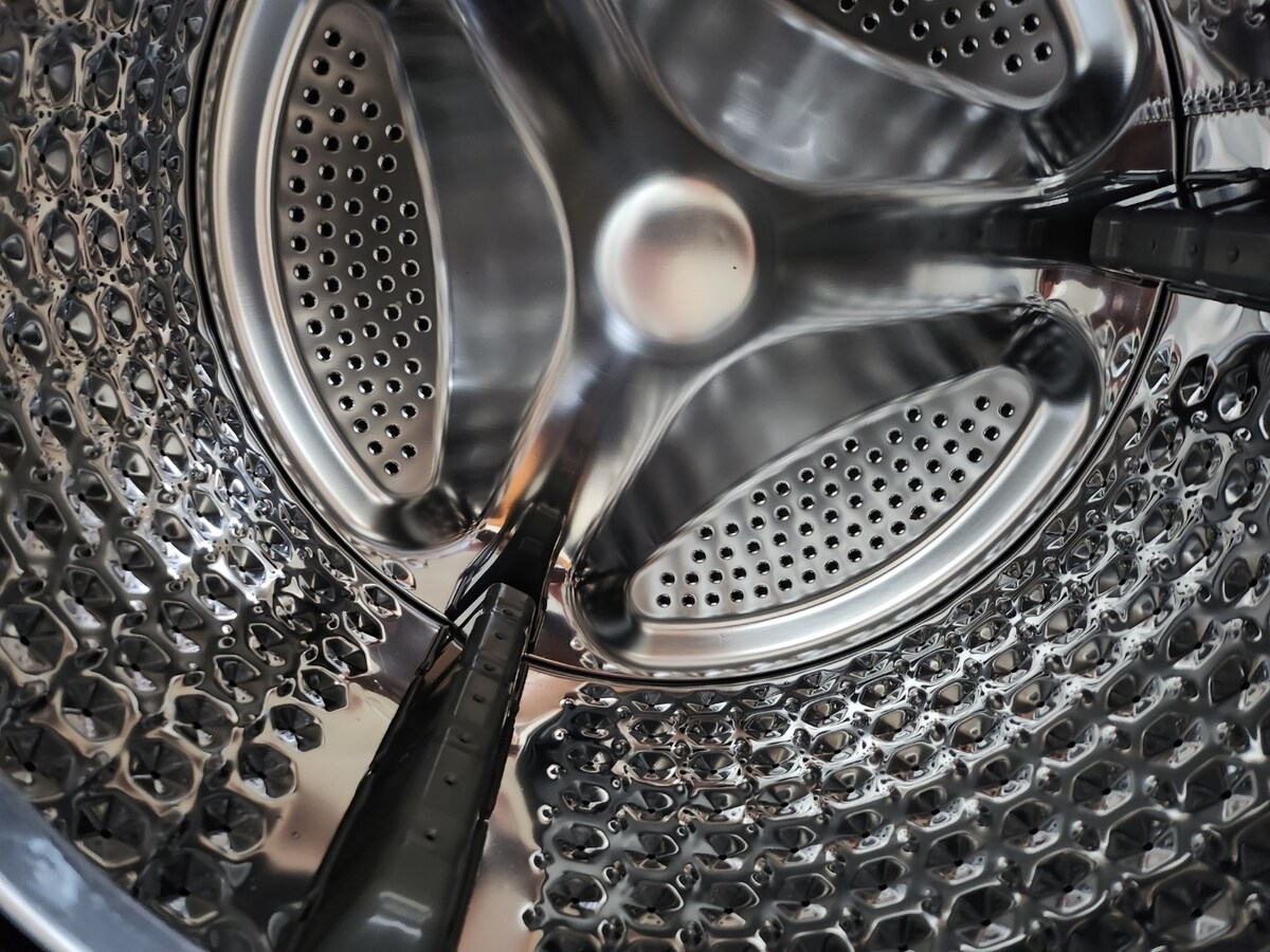 The interior of the washing machine drum is shown, featuring a shiny stainless steel surface with multiple perforations. The circular design allows for effective water drainage and is typical of standard washing machines.