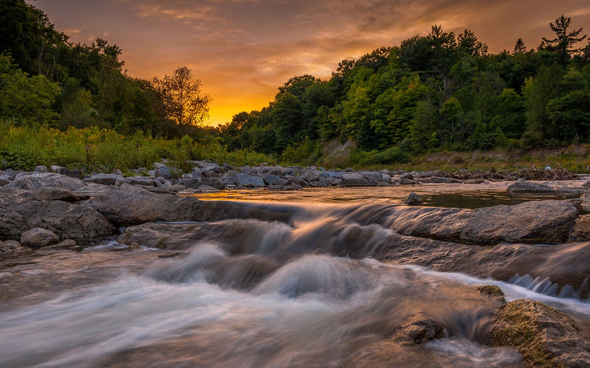 A serene river flows gently over smooth stones, reflecting the warm colors of a sunset. Lush greenery lines the banks, while the sky transitions from golden to deep orange, creating a tranquil setting. The landscape offers a peaceful connection to nature.