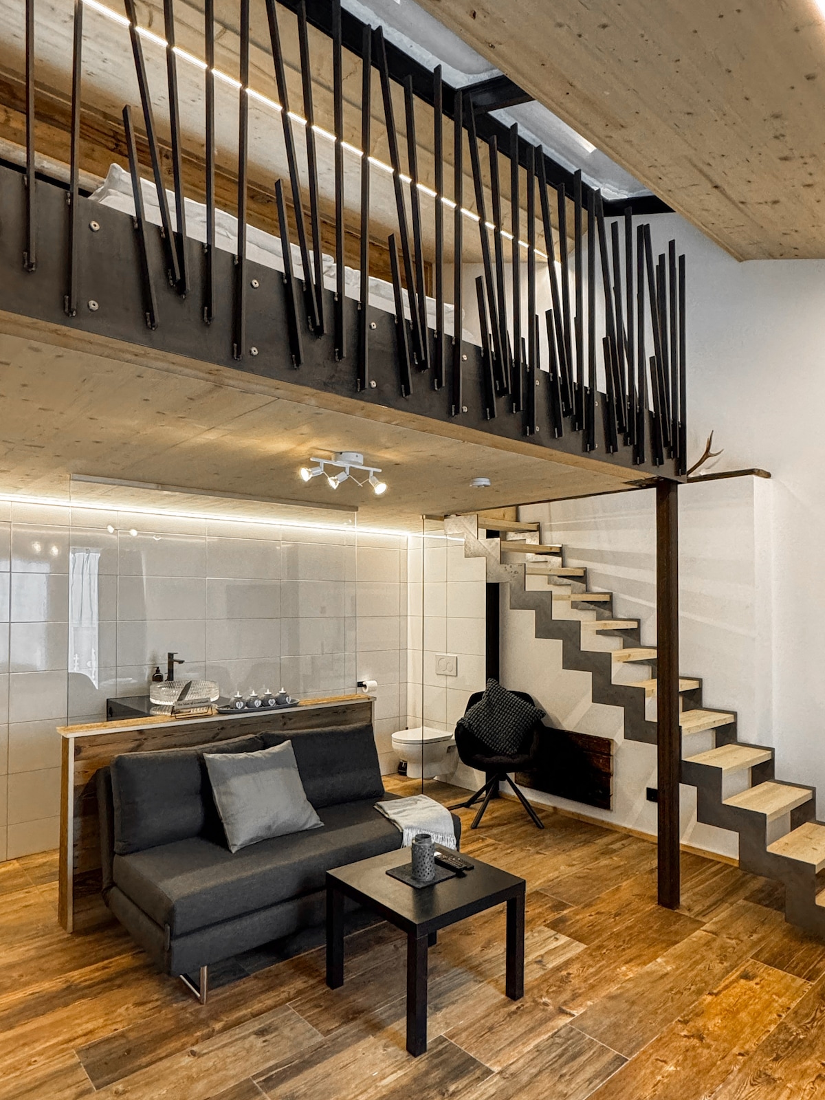 An open living area features a sleek black sofa paired with a minimalist black coffee table. A modern spiral staircase ascends to a lofted sleeping area, surrounded by distinctive vertical accents. Warm wooden flooring complements the clean lines and contemporary design of the space.