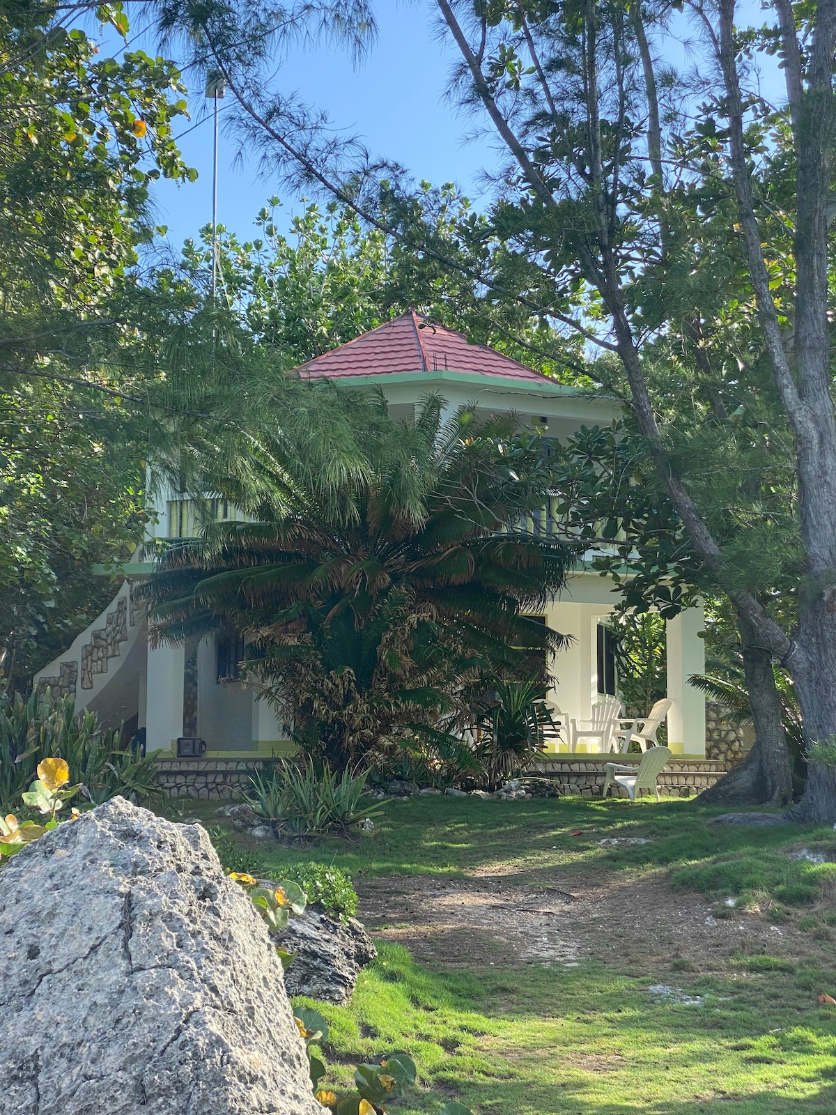 A two-story cottage appears amidst lush greenery, featuring a red-tiled roof and a balcony overlooking the surroundings. A staircase leads to the entrance, while a large palm tree and various plants frame the serene setting, creating a tranquil atmosphere.