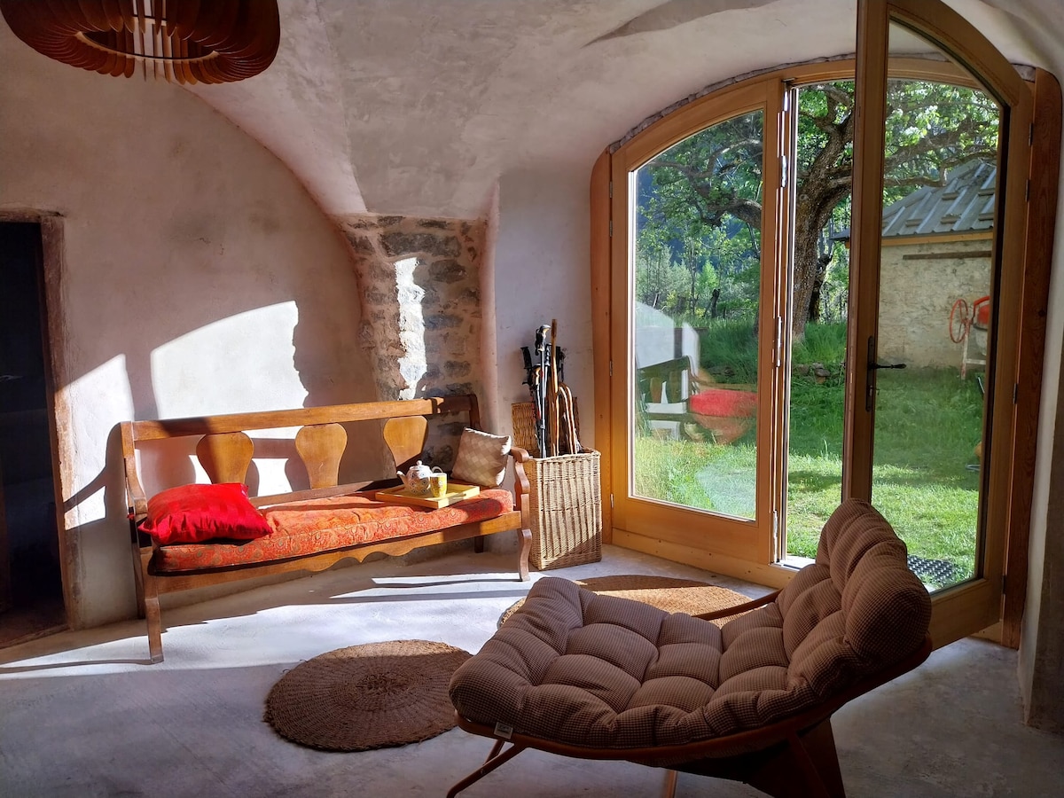 A sunlit living area features a rustic-style wooden bench with decorative cushions and a modern armchair. Large glass doors provide a view of the garden, while natural light enhances the earthy tones of the walls and floor.