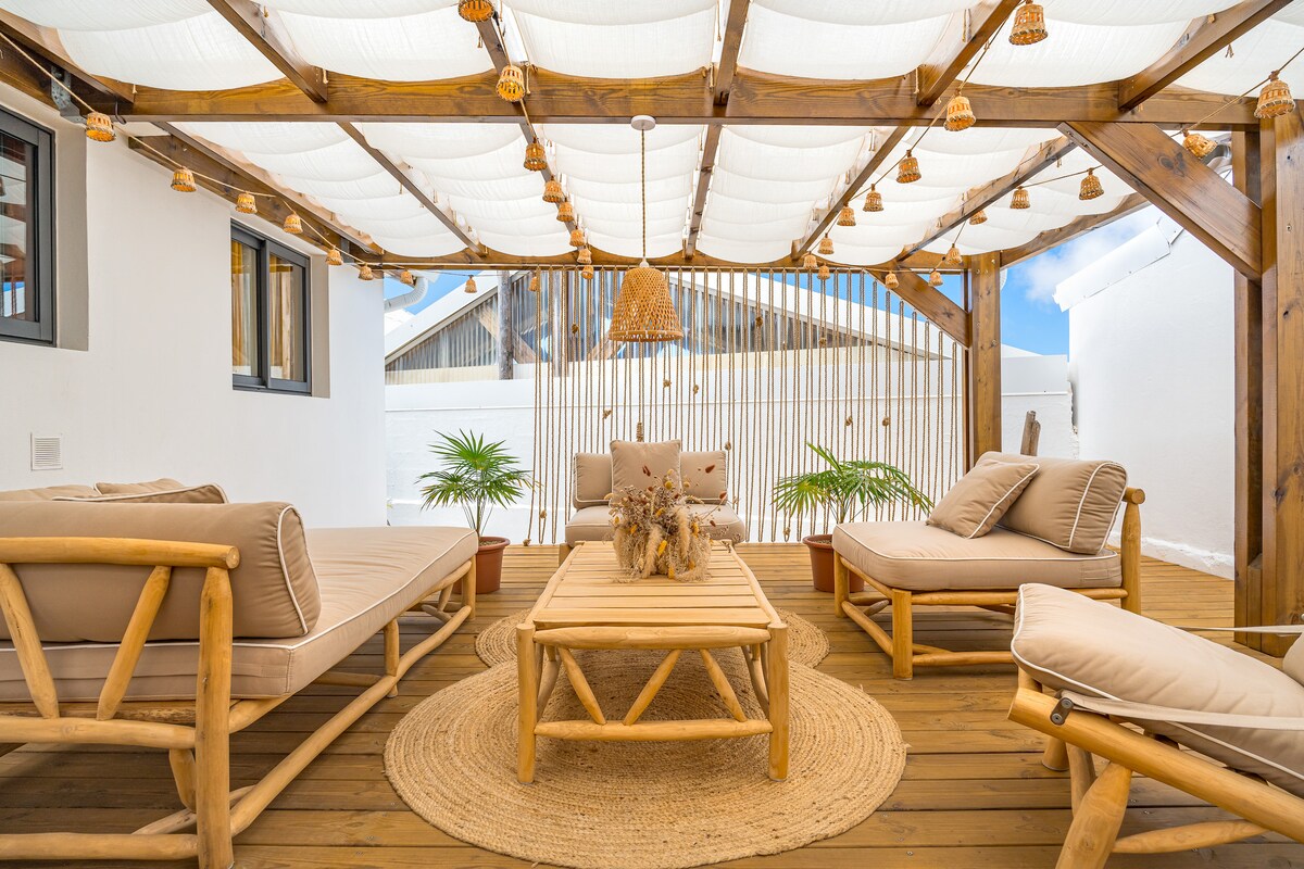 An outdoor terrace area features comfortable seating arrangements on wooden frames, complemented by a round woven rug. Soft cushions and a central wooden table create an inviting space. Overhead light fixtures and fabric canopies provide shade and a delightful atmosphere.