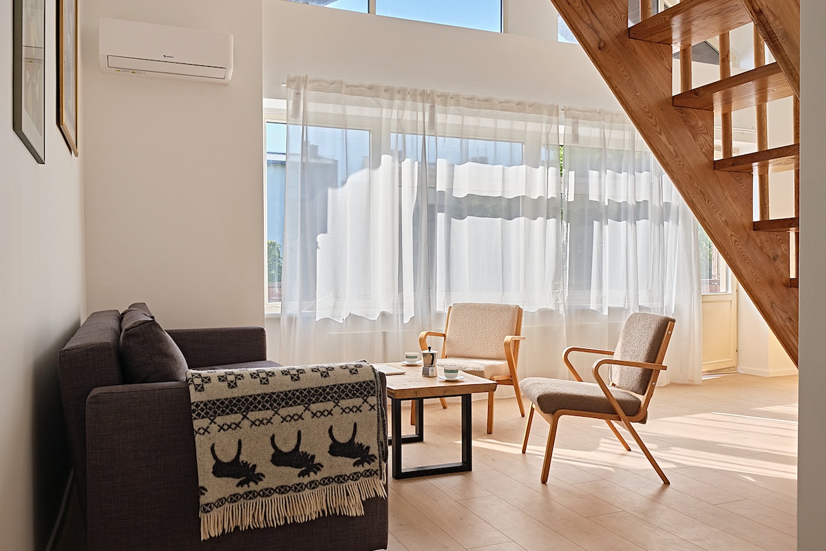 A well-lit living area features a comfortable sofa draped with a patterned throw, two armchairs, and a coffee table. Natural light streams in through large, sheer curtains, highlighting the warm wooden staircase in the background.