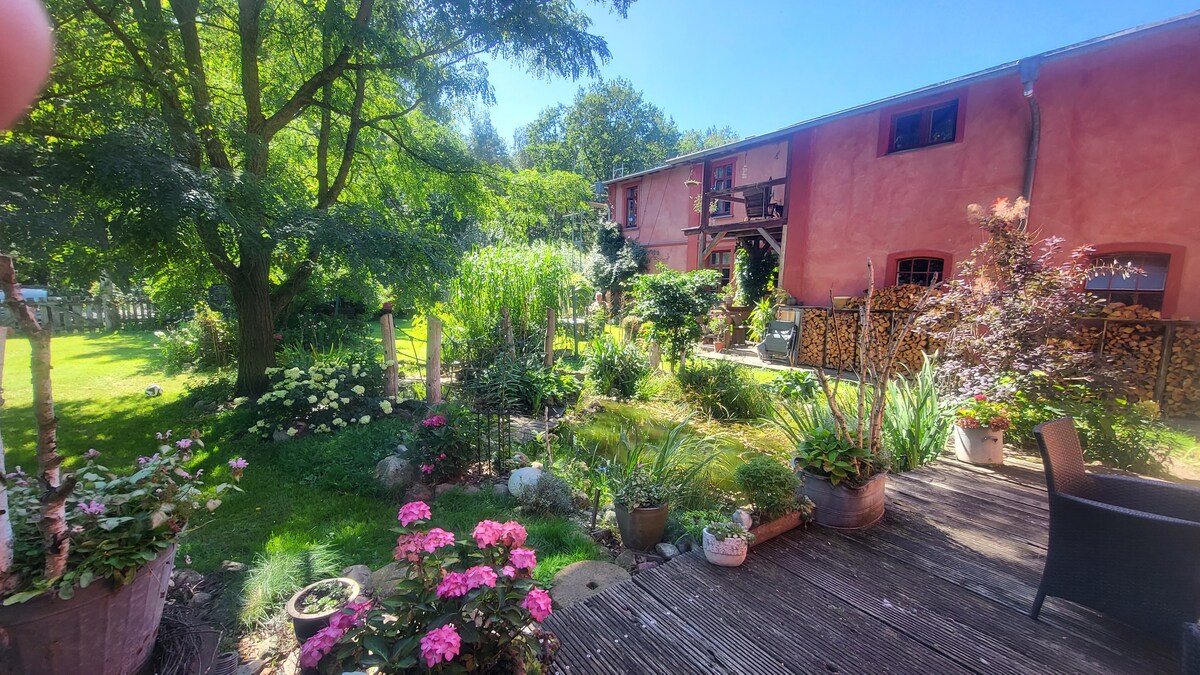The image features a garden area adorned with various potted plants, including vibrant flowers. A wooden deck extends into a grassy space surrounded by lush greenery and trees. The building in the background is painted in a soft hue, offering a quaint rural charm.