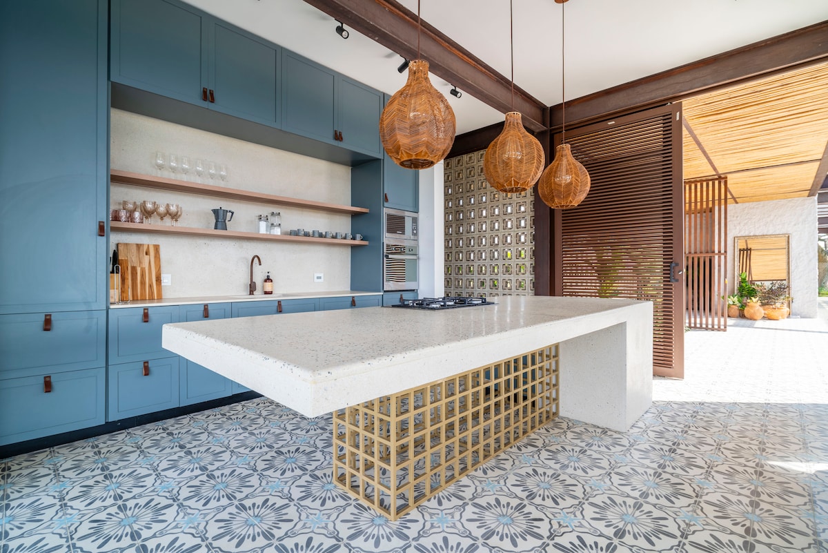 A modern kitchen is characterized by a large, textured island with a unique base made of woven materials. Three distinctive pendant lights hang overhead, complementing dark blue cabinetry and open shelving. Decorative tiles cover the floor, and natural light is invited through expansive openings.