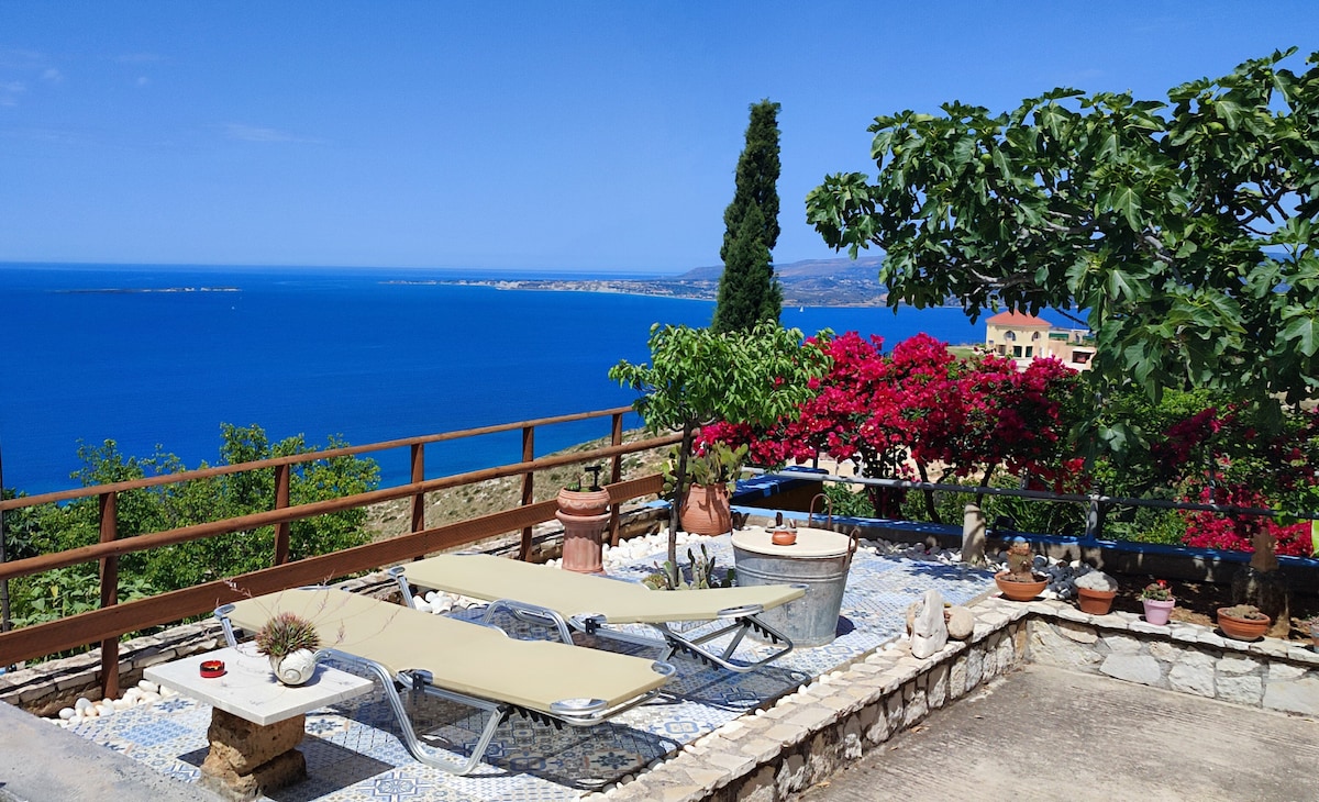 A sun-drenched terrace features two lounge chairs positioned to enjoy panoramic views of the Ionian Sea. Colorful flowers and greenery surround the space, enhancing the outdoor setting. The gentle slope of the coastline is visible in the background, complemented by the clear blue sky.