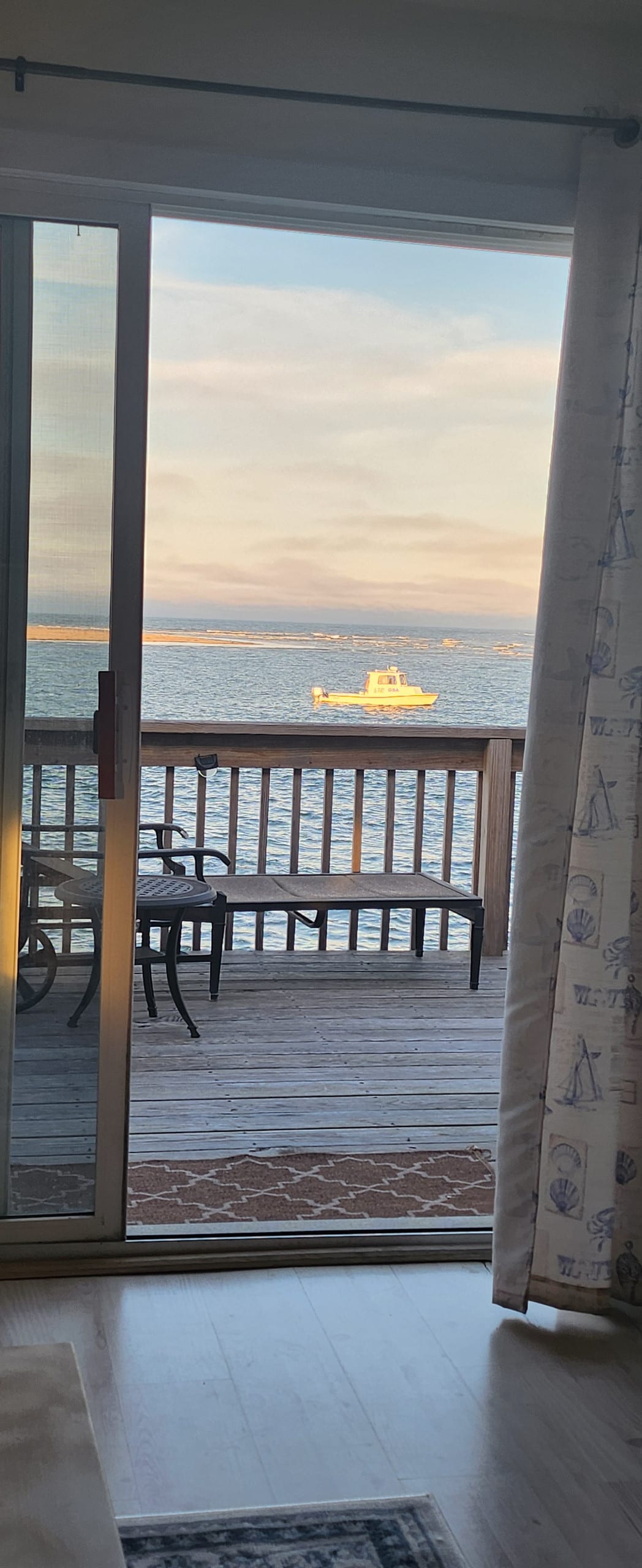 An open sliding glass door reveals a view of the ocean and a small boat navigating the waters. A wooden deck with a railing is visible, providing a tranquil outdoor space. Light from the setting sun casts a warm glow across the scene.