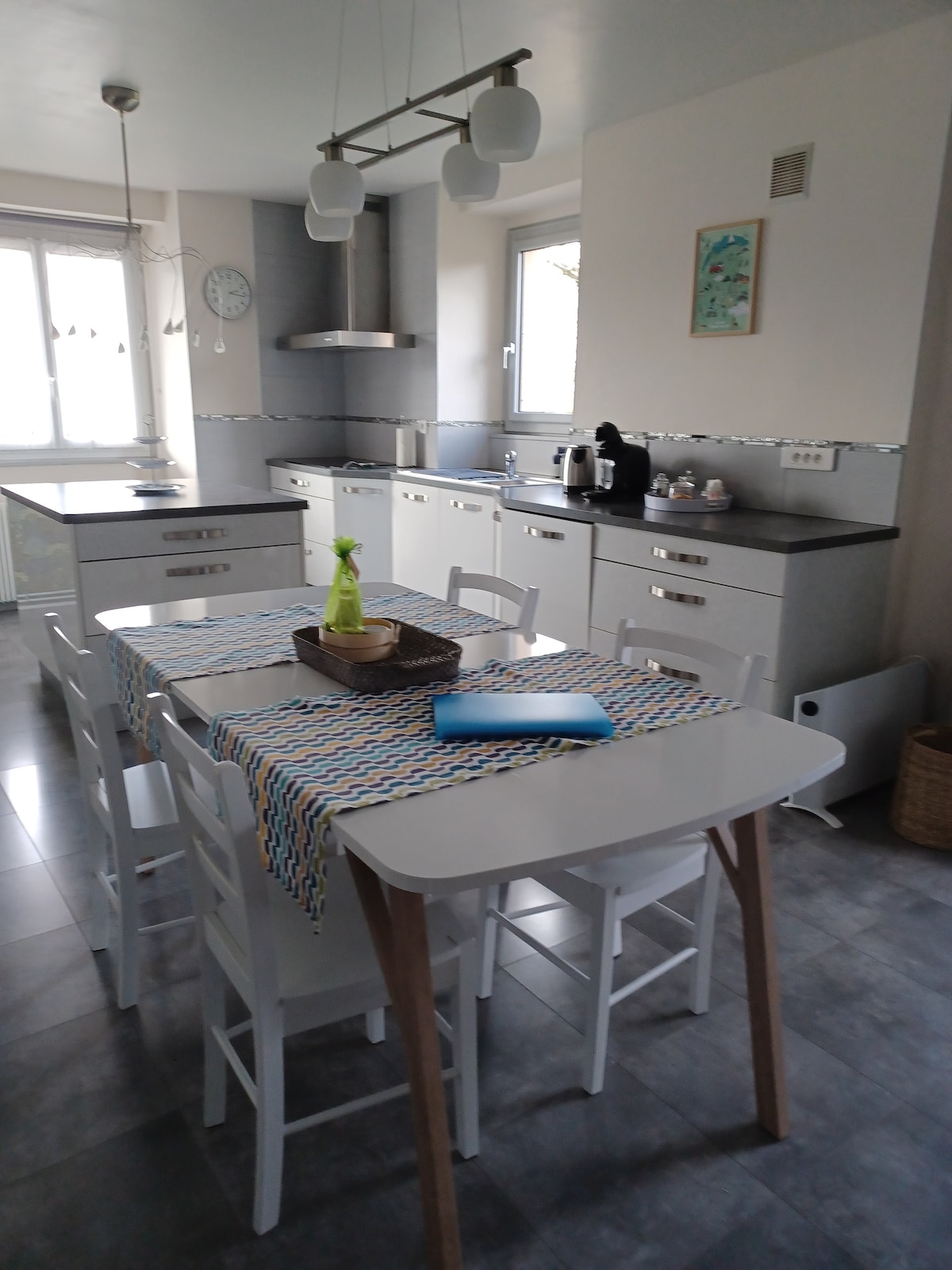 The kitchen features modern cabinetry and a central dining table surrounded by white chairs. A window allows natural light to enter, illuminating the countertop and appliances, including a kettle. Decorative items and tableware are arranged neatly, contributing to a functional and welcoming environment.