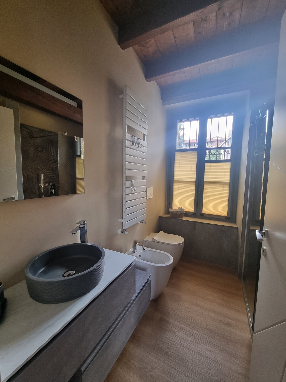 A modern bathroom is defined by clean lines and natural materials. A stylish sink sits atop a dark vanity, accompanied by a bidet and a toilet. Light enters through two windows, illuminating the space, while warm wooden beams add character to the high ceiling.