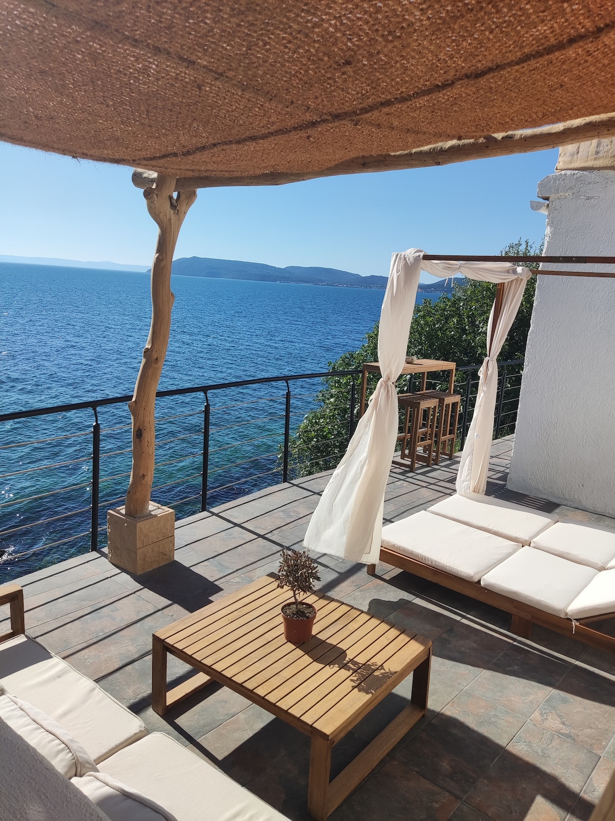 An outdoor terrace offers stunning views of the sea, featuring a wooden table and seating. Sunlight casts a warm glow over the space, while draped fabric adds a touch of elegance. The surrounding greenery enhances the tranquil setting, ideal for relaxation.