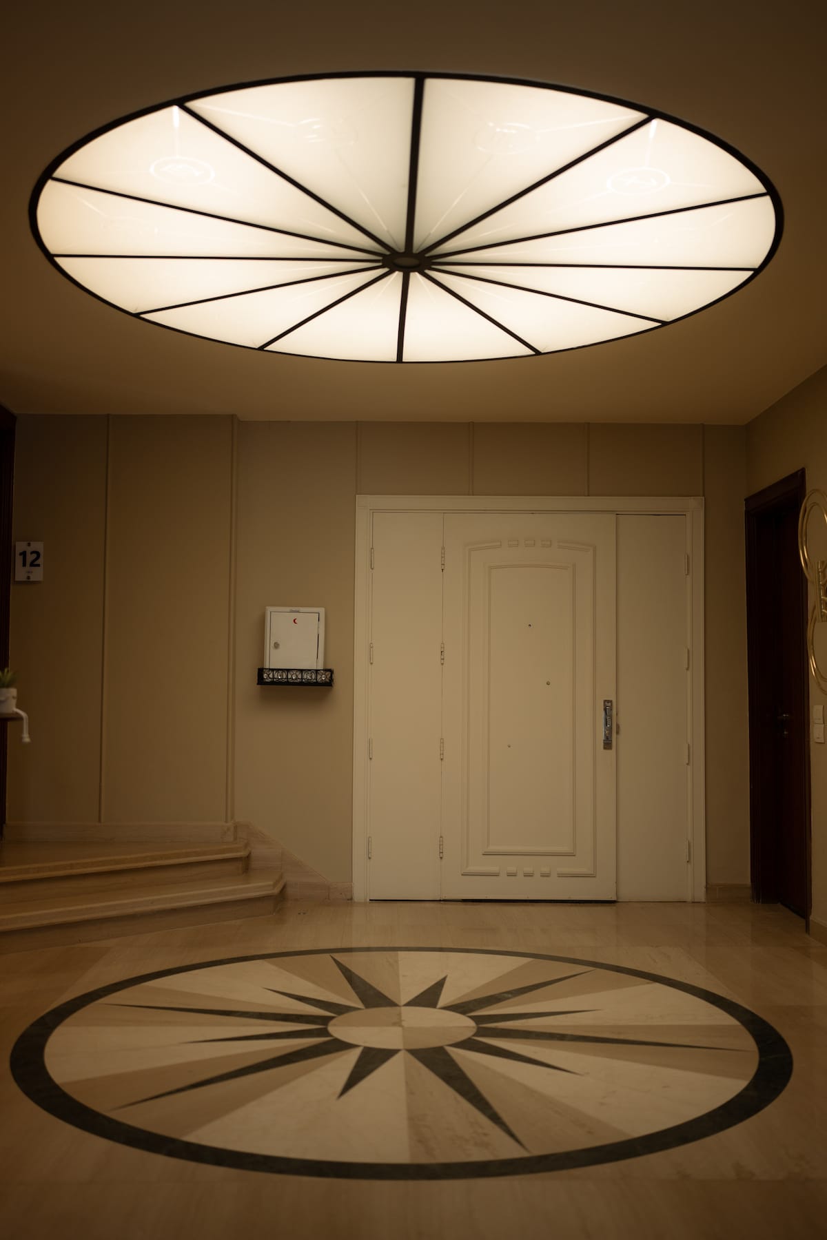 An elegant entryway is highlighted by a large, decorative ceiling fixture casting a soft glow. A detailed compass rose design is visible on the polished floor, while a simple white door is positioned prominently in the background.