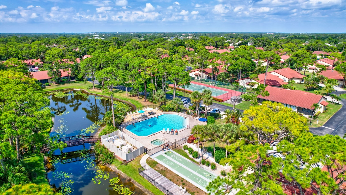 An aerial view showcases the resort-style community featuring a pool area surrounded by comfortable lounge chairs, tennis courts, and lush greenery. Calm ponds reflect the blue sky, enhancing the serene atmosphere of the landscape.