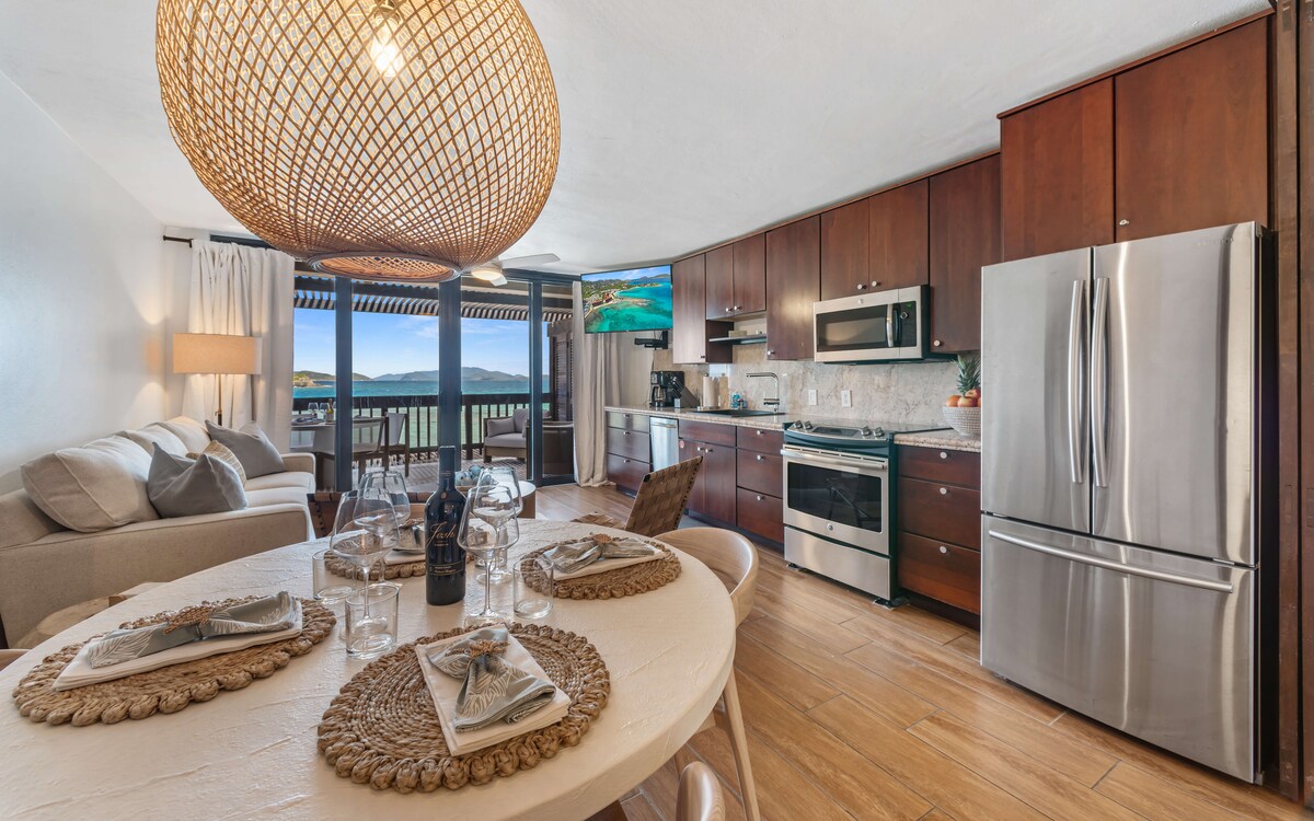 A modern kitchen features stainless steel appliances, including a refrigerator and oven. A round dining table, set with dinnerware, sits adjacent to a cozy living area with a neutral-colored sofa. Natural light floods the space through large glass doors leading to a balcony with scenic views.