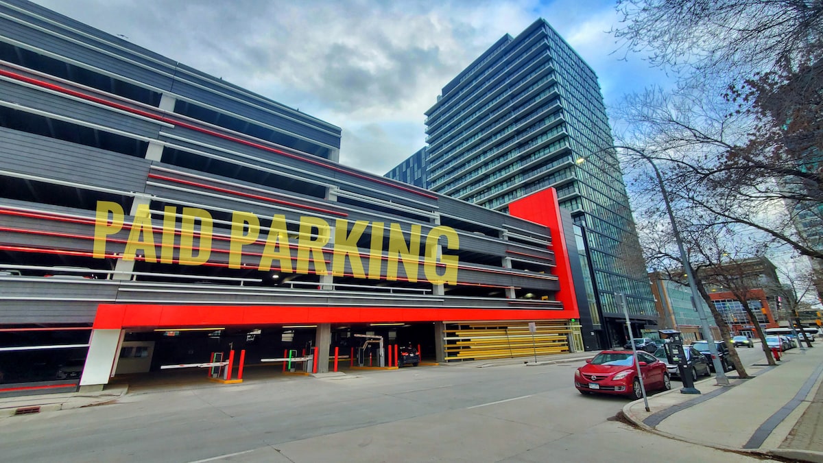 The image displays the exterior of a modern multi-story parking garage, characterized by a bold red accent and a sign indicating 'Paid Parking.' Adjacent buildings in the background contribute to the urban setting, with a tree-lined sidewalk visible in the foreground.