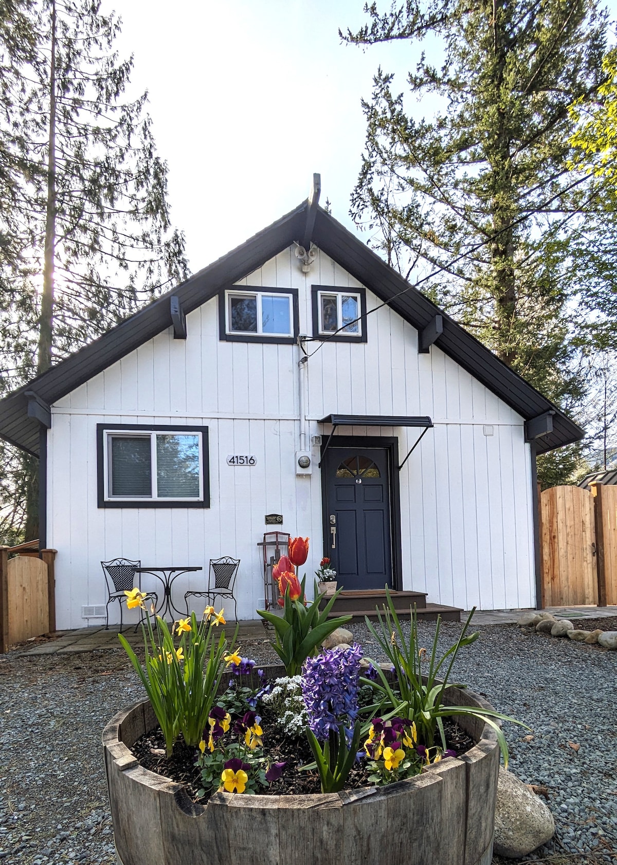 A charming cabin exterior is framed by tall trees, featuring a welcoming entrance with a dark blue door. A circular flower bed filled with vibrant spring flowers adds color to the gravel front yard, complemented by two outdoor chairs set nearby.