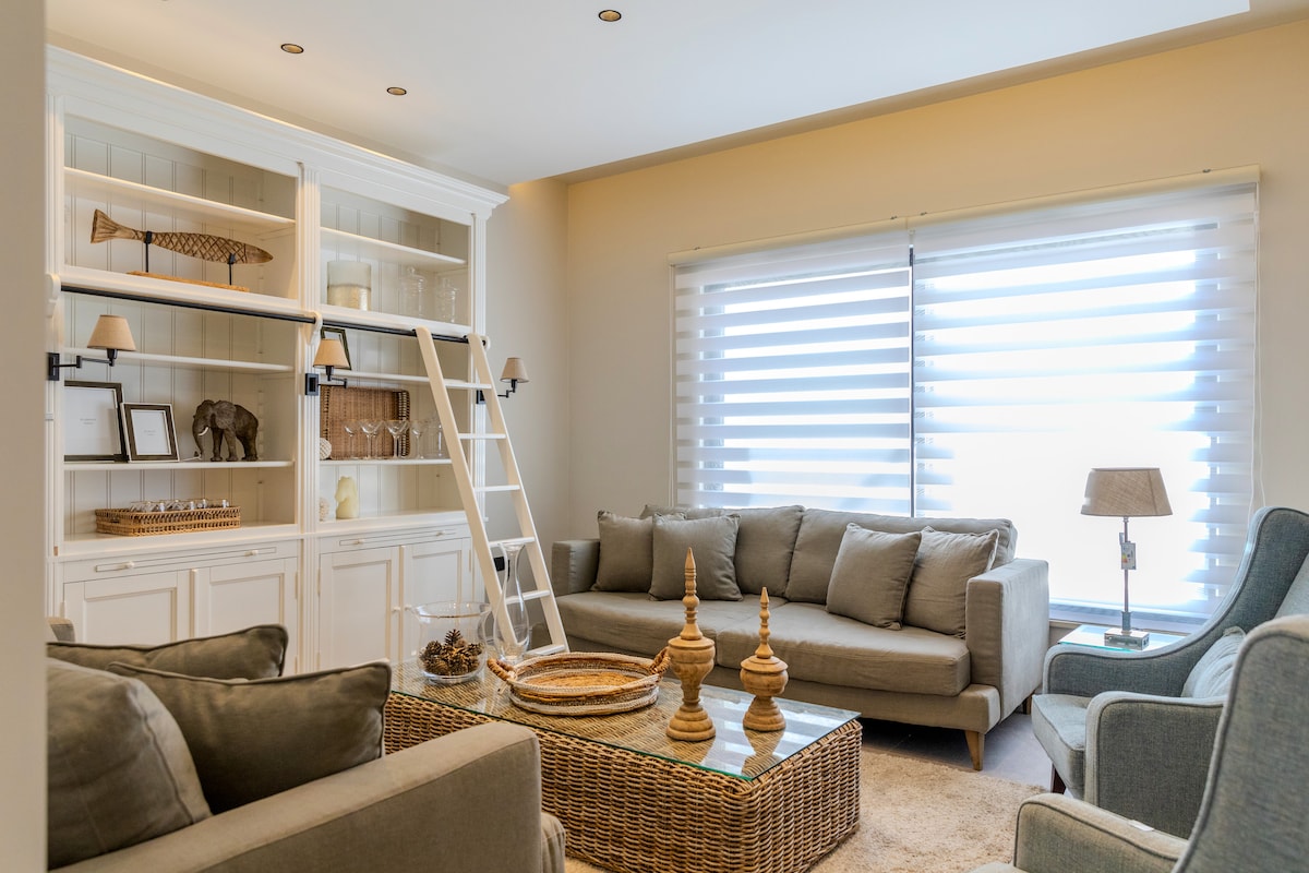 The spacious living area features a comfortable sectional sofa and a cozy armchair, complemented by a glass coffee table. A stylish shelving unit displays decorative items, while soft blinds filter natural light from the window. Warm tones invite a sense of relaxation.