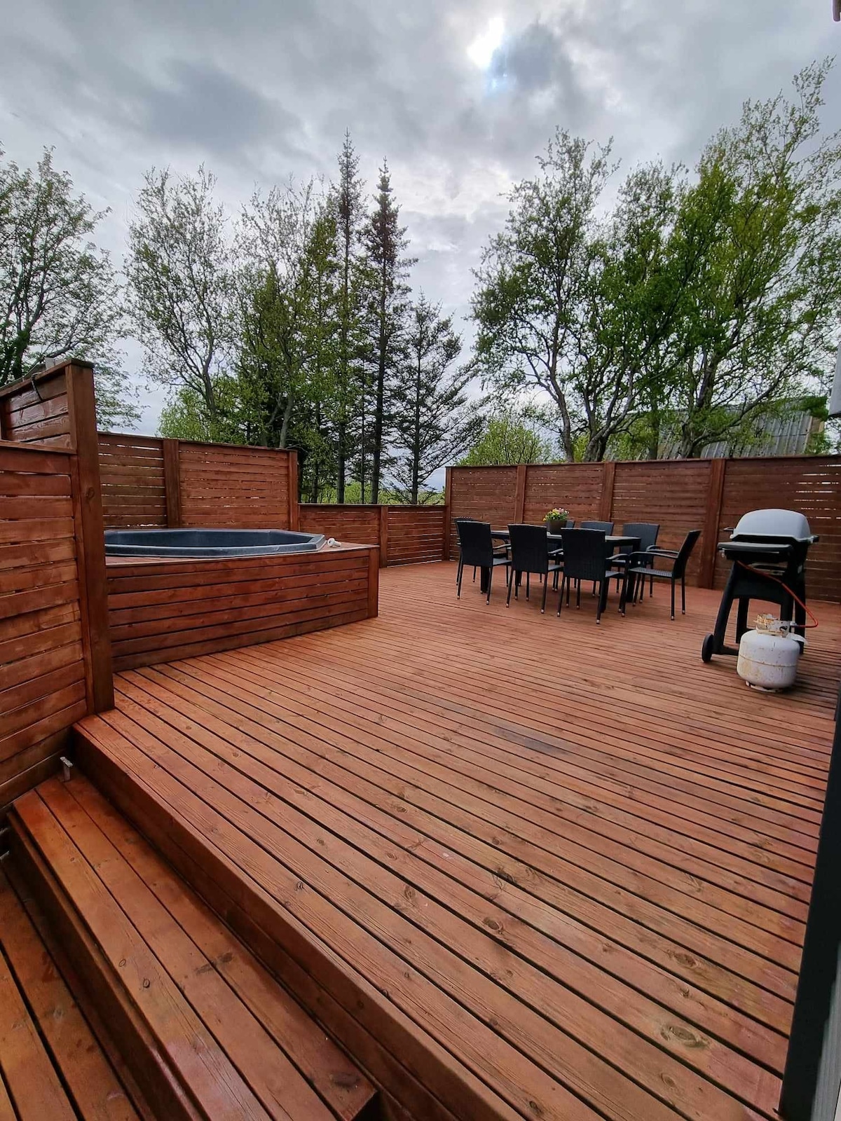 An outdoor deck is presented, featuring a hot tub and a dining table surrounded by chairs. A grill is positioned nearby. The wooden deck is bordered by wooden panels and is flanked by trees, providing a secluded outdoor environment.