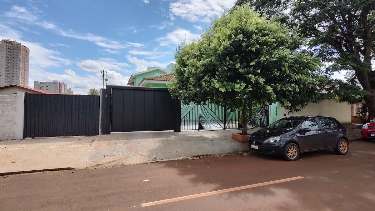 A view of the exterior of the property is presented, featuring a black gate with a green entrance nestled behind. A large tree provides shade beside a parked car, set against a backdrop of blue skies and distant buildings.