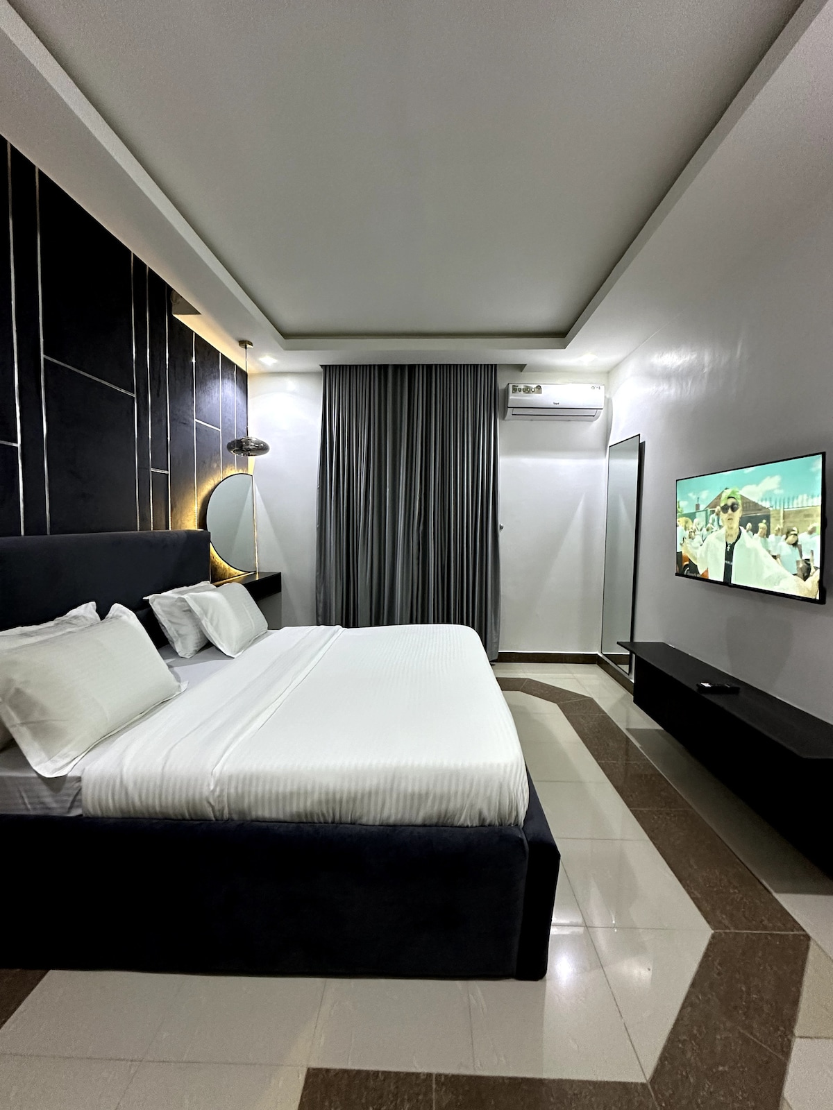 The bedroom features a spacious bed with crisp white linens and two pillows. A flat-screen TV is mounted on the wall opposite the bed. Soft lighting illuminates the room, with large curtains partially drawn to provide privacy.