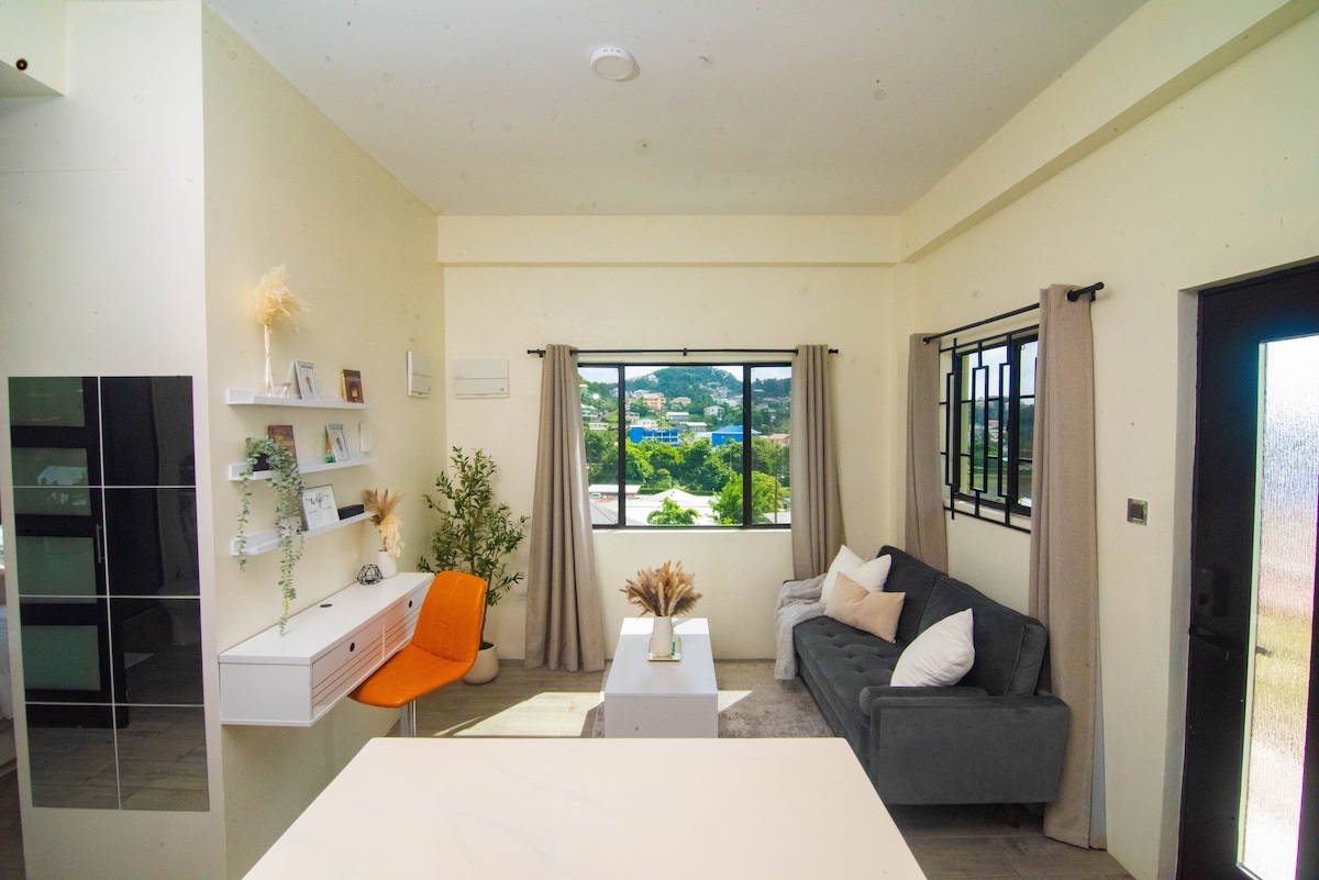 A modern studio space is presented, featuring a light-colored wall and a large window framing a view of greenery. A gray sofa and an orange chair are positioned beside a coffee table, while decorative plants add a touch of nature to the room.