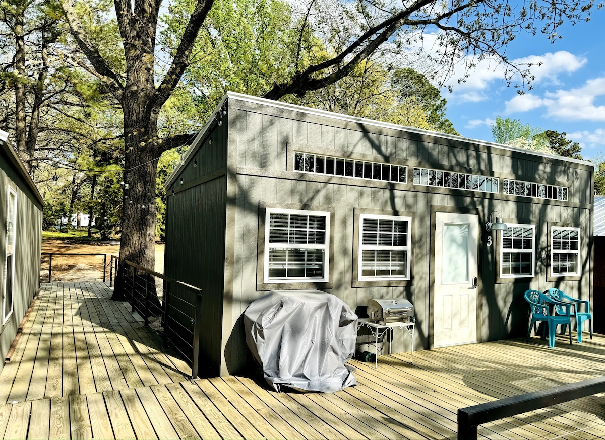 The exterior of a cozy cabin is shown, featuring a large wooden deck with a grill covered by a protective cover. Multiple windows allow natural light to enter, and colorful chairs are positioned nearby. Surrounding trees provide shade and greenery.