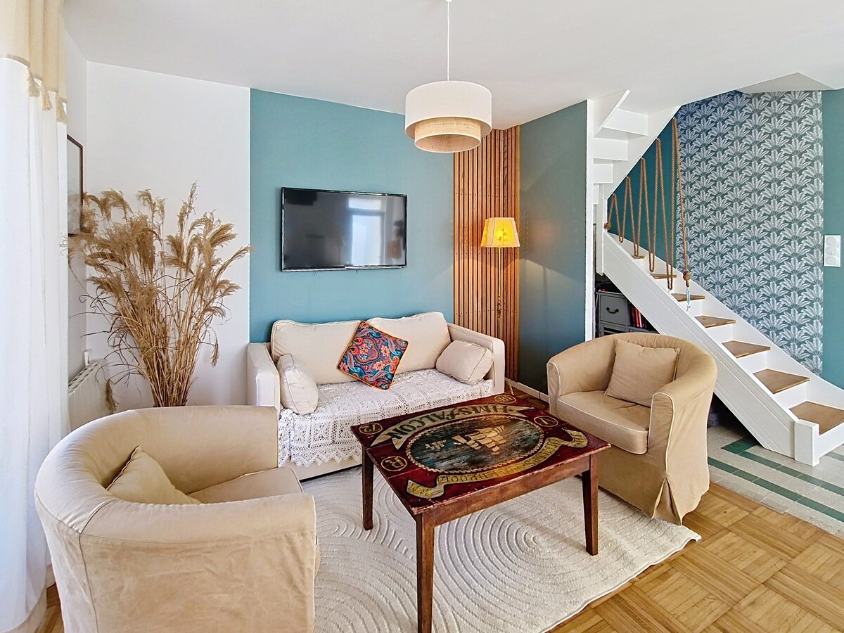 The living area features a light-colored sofa and two armchairs positioned around a wooden coffee table. A flat-screen television is mounted on the wall, while natural light filters through sheer curtains. A staircase with a wooden handrail leads to the upper level, complemented by decorative wall patterns.