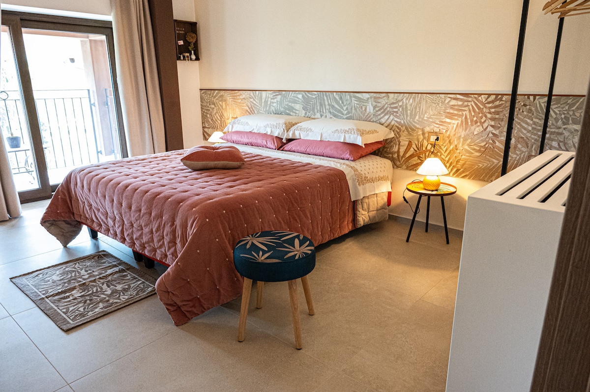 A spacious sleeping area is furnished with a plush, warmly colored bedspread covering a large bed, complemented by decorative pillows. A small, patterned stool sits nearby, and soft lighting is provided by bedside lamps. A private terrace can be seen through the open glass doors.